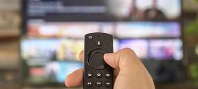 A hand holds an Amazon Fire TV Stick Lite remote toward a TV